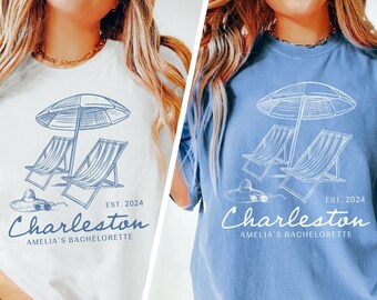 May include: White and blue jean t-shirts with a graphic of beach chairs and an umbrella. The text "Charleston" and "Amelia's Bachelorette" is printed on the shirts. EST. 2024 is also printed on the shirts.