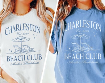 May include: Two women's t-shirts with a graphic design of beach chairs and an umbrella. The text on the shirts reads "Charleston Est. 2024 Beach Club Amelia's Bachelorette". The shirts are white and blue jean.