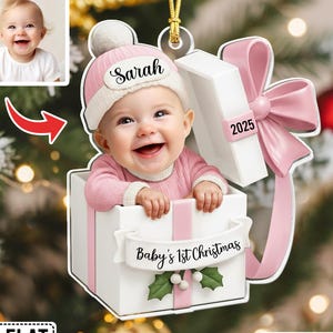 May include: A personalized baby's first Christmas ornament featuring a smiling baby in a pink outfit and hat, emerging from a white gift box with a pink bow. The ornament includes the name "Sarah" and the year "2025". The banner reads "Baby's 1st Christmas".