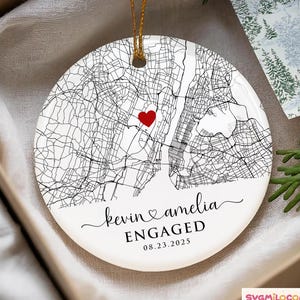 May include: A white ceramic ornament with a map of a city, a red heart, and the words "Kevin & Amelia ENGAGED 08.23.2025". The ornament has a gold string for hanging. The ornament is in a gift box.