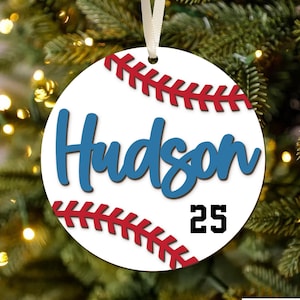 May include: A white baseball ornament with red stitching and the name 'Hudson' in blue script. Below the name is the number '25' in black. The ornament is round and has a white ribbon for hanging. The background is a blurred Christmas tree.