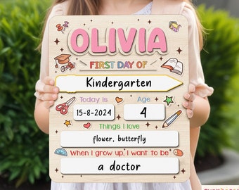 May include: A wooden sign with a pink background and the name Olivia written in white. The sign says "First Day of Kindergarten" and includes a date, age, and space to write about things the child loves and what they want to be when they grow up.