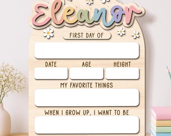 Puede incluir: Un letrero de madera arqueado con el nombre Eleanor en letra cursiva colorida, decorado con margaritas blancas. El tablero incluye espacios en blanco para registrar detalles del primer día de clases como fecha, edad, altura, favoritos y metas.