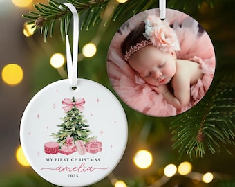 May include: Two round Christmas ornaments hanging from a tree. One features a baby photo, the other has a Christmas tree design with gifts and the text "My First Christmas Amelia 2025". Both have white ribbons.