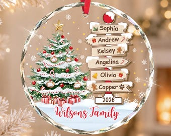 May include: A round, clear glass Christmas ornament featuring a decorated pine tree with red ornaments and gifts, a signpost with the names Sophia, Andrew, Kelsey, Angelina, Olivia, and Copper, the year 2026, and the text Wilsons Family in red script.