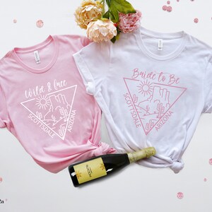 May include: Two white t-shirts with pink text and a graphic of a desert landscape. The text on one shirt says "Wild & Free" and the text on the other shirt says "Bride to Be". Both shirts have the words "Scottsdale" and "Arizona" on them.