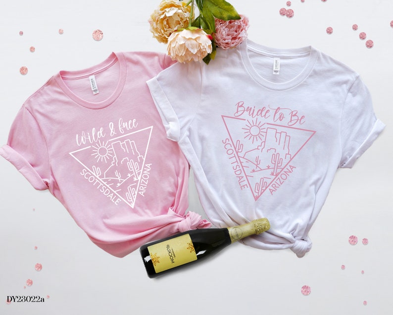 May include: Two white t-shirts with pink text and a graphic of a desert landscape. The text on one shirt says "Wild & Free" and the text on the other shirt says "Bride to Be". Both shirts have the words "Scottsdale" and "Arizona" on them.