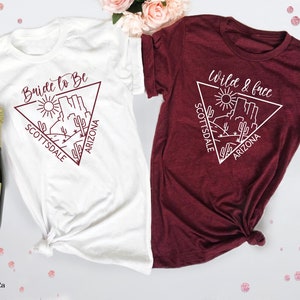 May include: Two white t-shirts with a maroon design. The left shirt says "Bride to Be" with a triangle design featuring a desert scene and the words "Scottsdale Arizona". The right shirt says "Wild & Free" with a triangle design featuring a desert scene and the words "Scottsdale Arizona".