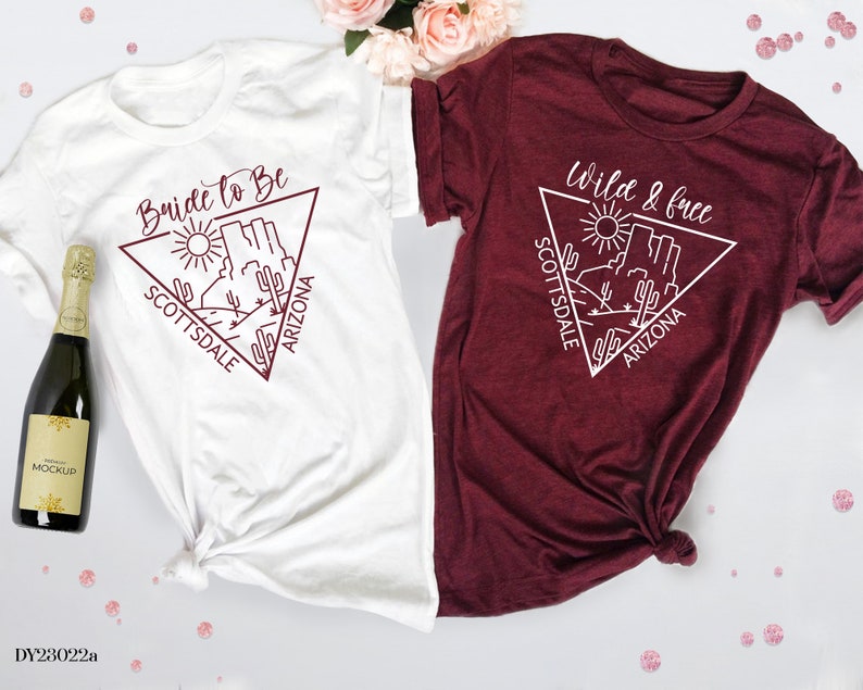 May include: Two white t-shirts with a maroon design. The left shirt says "Bride to Be" with a triangle design featuring a desert scene and the words "Scottsdale Arizona". The right shirt says "Wild & Free" with a triangle design featuring a desert scene and the words "Scottsdale Arizona".