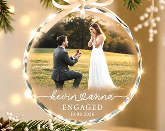 May include: A circular glass ornament featuring a photograph of a proposal in a field. The text reads "kevin & anna", "ENGAGED", and "10.08.2026". The ornament is topped with a cream-colored ribbon bow and has a faceted, reflective edge.