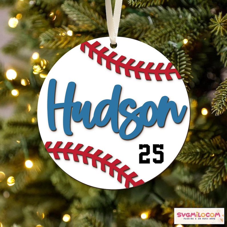 May include: A white baseball ornament with red stitching and the name 'Hudson' in blue script. Below the name is the number '25' in black. The ornament is round and has a white ribbon for hanging. The background is a blurred Christmas tree.