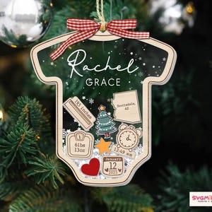 May include: A baby onesie-shaped Christmas ornament made of wood with a dark green background featuring white snowflakes. It displays the name Rachel Grace, a ruler labeled 195 inches, a map of Arizona, a tree, a scale reading 61lbs 13oz, a clock, a star, a heart, and a calendar date of January 12 2025, topped with a red and white gingham ribbon.