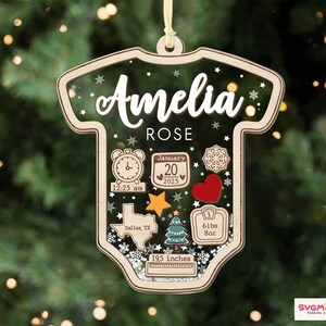May include: A baby bodysuit-shaped wooden ornament featuring the name Amelia Rose, birth date January 20, 2025, time 12:25 am, location Dallas, TX, weight 61 lbs 8 oz, and length 19.5 inches, decorated with a clock, heart, star, and tree.