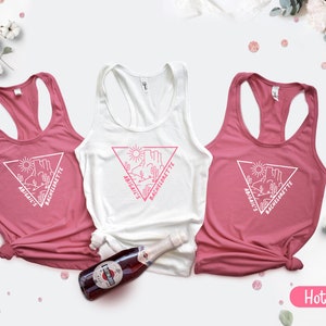 May include: Three pink tank tops with white text that says "Abigail's Bachelorette" and a graphic of a desert scene with a triangle around it. The tank tops are on a white background with a bottle of sparkling wine.