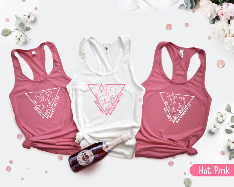 May include: Three pink tank tops with white text that says "Abigail's Bachelorette" and a graphic of a desert scene with a triangle around it. The tank tops are on a white background with a bottle of sparkling wine.