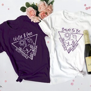 May include: Two white t-shirts with a purple triangle design featuring a desert landscape and the text "Bride to Be Scottsdale Arizona" and "Wild & Free Scottsdale Arizona".