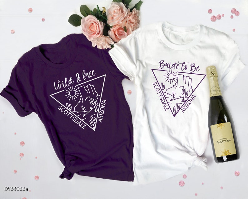 May include: Two white t-shirts with a purple triangle design featuring a desert landscape and the text "Bride to Be Scottsdale Arizona" and "Wild & Free Scottsdale Arizona".