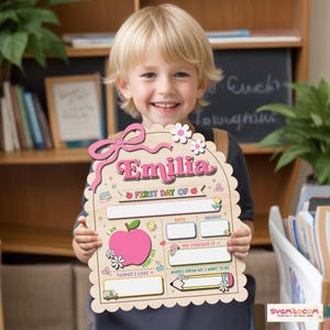 Peut inclure: Une plaque en bois clair &agrave; bord festonn&eacute; portant le pr&eacute;nom Emilia en lettres roses. La plaque comprend des sections pour First Day Of, Date, Height, My Teacher Is, Things I Love et When I Grow Up, I Want To Be, avec des illustrations.