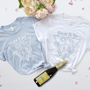 May include: Two light blue and white t-shirts with a graphic design of a desert landscape with the text "Wild & Free Scottsdale Arizona" and "Bride to Be Scottsdale Arizona".