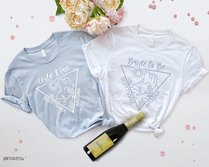 May include: Two light blue and white t-shirts with a graphic design of a desert landscape with the text "Wild & Free Scottsdale Arizona" and "Bride to Be Scottsdale Arizona".