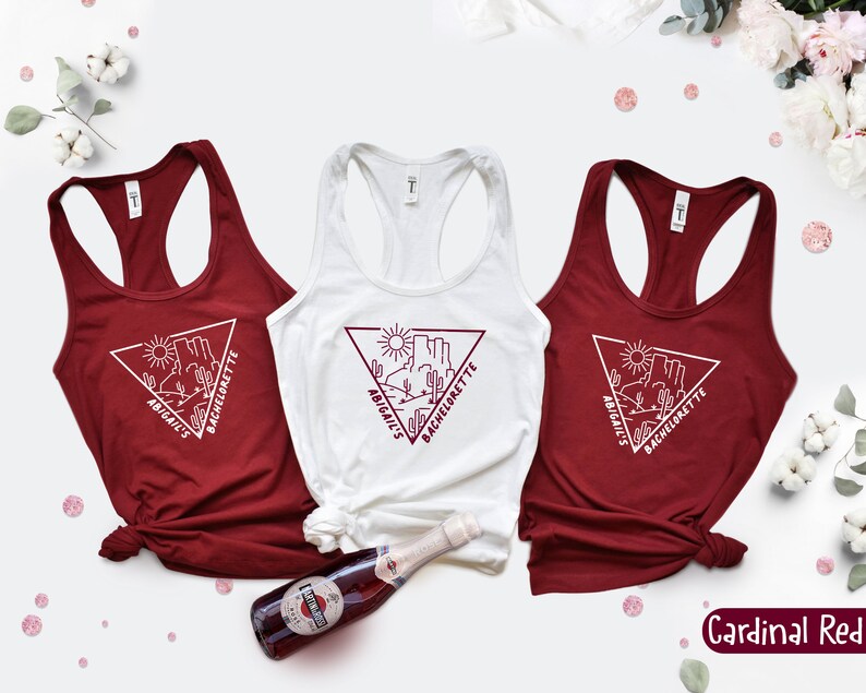 May include: Three red tank tops with white text and a graphic of a desert scene with the words "Abigail's Bachelorette" on each. The tank tops are on a white background with a bottle of sparkling wine and pink confetti.