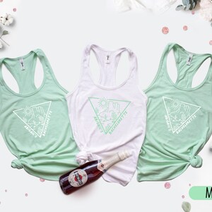 May include: Three mint green tank tops with a white triangle graphic and the text "Abigail's Bachelorette" on each. The tank tops are laid out on a white surface with a bottle of sparkling wine and pink confetti.