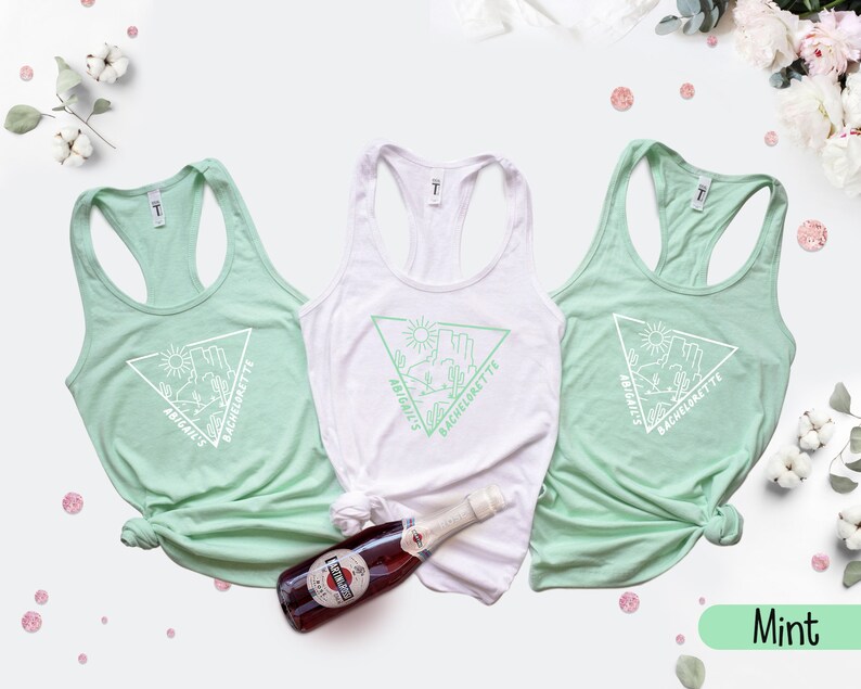May include: Three mint green tank tops with a white triangle graphic and the text "Abigail's Bachelorette" on each. The tank tops are laid out on a white surface with a bottle of sparkling wine and pink confetti.