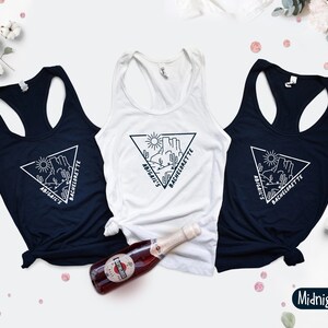 May include: Three white tank tops with black text and a graphic of a desert landscape in a triangle. The text reads "Abigail's Bachelorette". The tank tops are on a white background. One tank top is midnight navy.