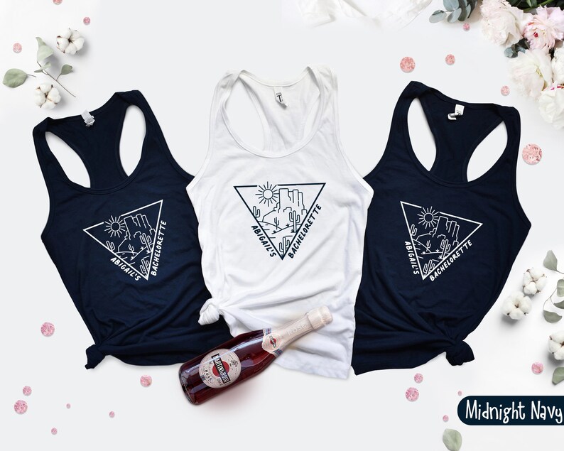 May include: Three white tank tops with black text and a graphic of a desert landscape in a triangle. The text reads "Abigail's Bachelorette". The tank tops are on a white background. One tank top is midnight navy.