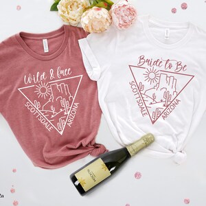 May include: Two white t-shirts with a graphic design of a desert landscape in a triangle with the text "Wild & Free" and "Bride to Be" in pink. The text "Scottsdale Arizona" is also included in the design.