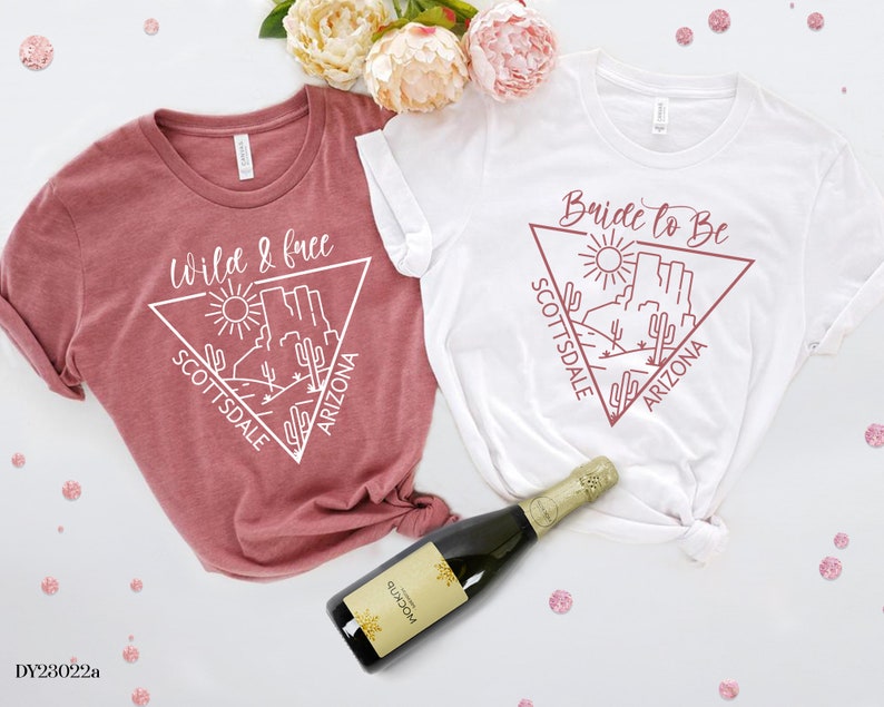 May include: Two white t-shirts with a graphic design of a desert landscape in a triangle with the text "Wild & Free" and "Bride to Be" in pink. The text "Scottsdale Arizona" is also included in the design.