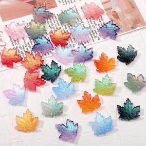 Può includere: Una collezione di abbellimenti a forma di foglia d'acero colorati in varie tonalità, tra cui arancione, verde, blu e viola. Le foglie hanno un aspetto glitterato e traslucido. Il testo "Medusa BoutiqueStore" è visibile in alto.