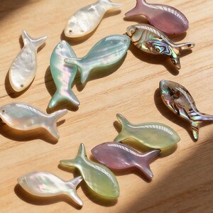 May include: Various iridescent fish-shaped beads in shades of white, pink, green, and abalone, scattered on a wooden surface. The text "Medusa Boutique Store" is at the top. These beads could be used for jewellery making.