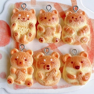 May include: Six orange and yellow bear-shaped charms with silver-colored loops. Some bears hold fish. The charms have a textured appearance and are likely made of resin. The text "Medusa BoutiqueStore" is at the top.