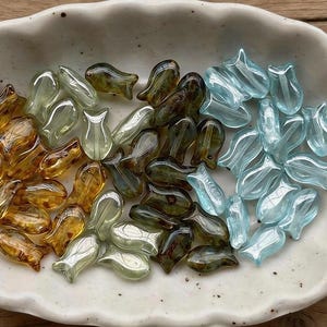 May include: Translucent glass beads in the shape of tulips, in amber, olive green, and light blue. The beads are arranged in a white ceramic dish. The shop name "Medusa Boutique Store" is at the top.