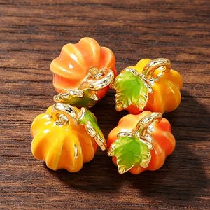 Op de afbeelding: Vier kleine pompoenvormige bedels met glanzend oranje emaille, goudkleurige metalen steeltjes en felgroene blaadjes. Elke bedel heeft een klein goudkleurig metalen oogje aan de bovenkant voor bevestiging, met een gladde, reflecterende afwerking.