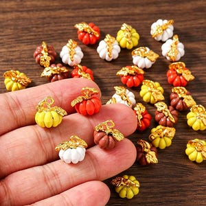 May include: A collection of small pumpkin-shaped charms in white, yellow, orange, and brown, each featuring a metallic gold stem and leaf detail. These miniature decorative pieces are scattered across a surface, with several held in a hand.