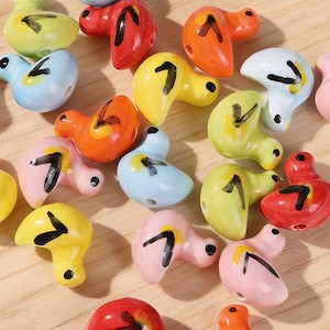 May include: A collection of colorful ceramic duck beads in various shades including yellow, red, orange, blue, and pink. Each bead features a black arrow design and a small hole for stringing. The beads are scattered on a light brown surface.