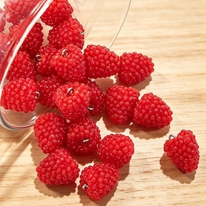 May include: A collection of bright red raspberry-shaped charms spilling from a clear glass container. Each individual charm features a textured, bumpy surface and a small silver-toned metal loop at the top for attachment. The text MedusaBoutiqueStore is visible at the top.