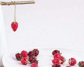 Può includere: Una collana con catena dorata con un ciondolo a forma di fragola rossa è appesa sopra un piatto bianco che contiene diversi ciondoli a forma di fragola rossa. I ciondoli hanno dettagli dorati e sono sparsi sul piatto. L'immagine ha il testo "Medusa Boutique Store" in alto.