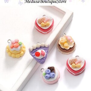 May include: A collection of miniature cake charms in various shapes and colors. The charms include heart-shaped, round, and triangular designs, each adorned with colorful frosting and toppings. The charms are made of resin and have a silver loop for attaching to jewelry.