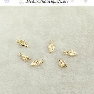 May include: Six small, gold-coloured leaf charms with a loop at the top for jewellery making. The charms are scattered on a white, textured surface. The text "Medusa Boutique Store" is at the top.
