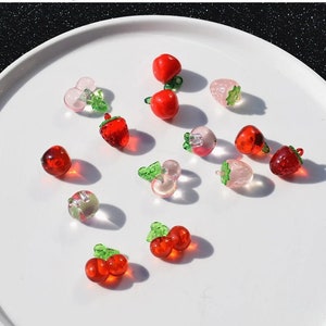 May include: Assortment of red and pink glass fruit charms, including strawberries and cherries, with green leaves.