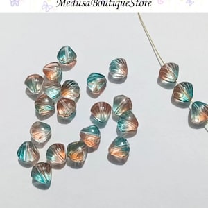 May include: A scattering of teal and peach colored glass beads shaped like seashells. The beads are arranged on a white surface.