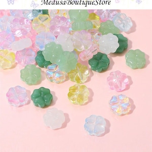 May include: Assortment of colourful four-leaf clover shaped beads. The beads are made of glass and have a variety of iridescent finishes. The beads are scattered on a pink surface.