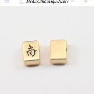 Puede incluir: Dos cuentas rectangulares doradas. Una cuenta tiene un car&aacute;cter chino negro. La otra cuenta es lisa. Las cuentas son probablemente para la fabricaci&oacute;n de joyas. La parte superior de la imagen tiene el texto "Medusa Boutique Store".