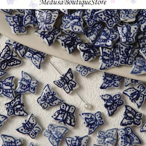 Può includere: Una collezione di piccole perline a farfalla in porcellana bianca con intricati motivi blu. Le perline sono sparse su una superficie bianca, con uno spillo e una perla visibili. Il testo "Medusa Boutique Store" è in alto.