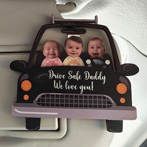 May include: A car-shaped ornament featuring three babies in the front seats. The black car has orange headlights and a silver grill. The text "Drive Safe Daddy We love you!" is written in white script.