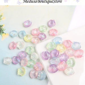 May include: A selection of iridescent seashell-shaped beads in pastel shades of pink, purple, blue, and yellow. The beads are scattered on a light blue surface and a white surface. The text "Medusa Boutique Store" is at the top.
