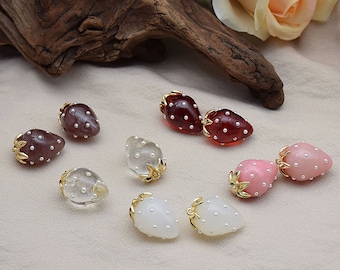 Può includere: Assortimento di orecchini in resina a forma di fragola in vari colori, tra cui rosso, rosa, bianco e trasparente. Ogni orecchino presenta una montatura in metallo dorato con un design a foglia e piccoli punti bianchi.